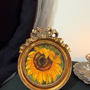 May include: An oval-shaped gold-toned frame with a decorative bow at the top, containing an oil painting of a vibrant yellow sunflower with a dark brown center. The frame features a beaded inner edge and a smooth, polished metallic finish.