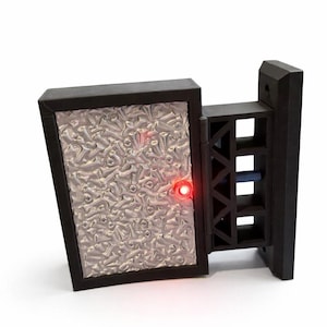 以下が含まれることがあります： テクスチャ加工された銀色の表面と赤いライトを備えた黒い長方形のデバイス。デバイスは、一連の正方形の開口部を備えた黒い支持構造に取り付けられています。背景は白です。