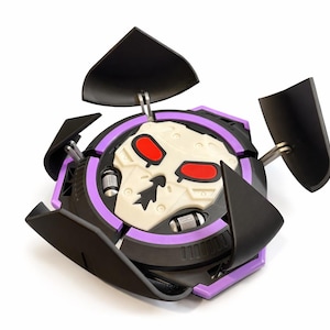 Puede incluir: Un juguete negro y morado con un dise&ntilde;o de calavera. El juguete tiene forma circular con una calavera blanca en el centro, ojos rojos y detalles negros. Tiene cuatro paneles negros que se pueden abrir.