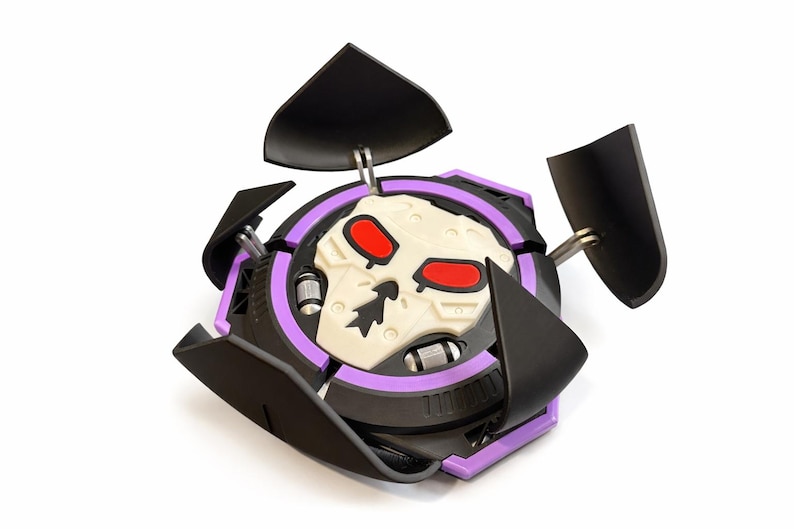 Puede incluir: Un juguete negro y morado con un dise&ntilde;o de calavera. El juguete tiene forma circular con una calavera blanca en el centro, ojos rojos y detalles negros. Tiene cuatro paneles negros que se pueden abrir.