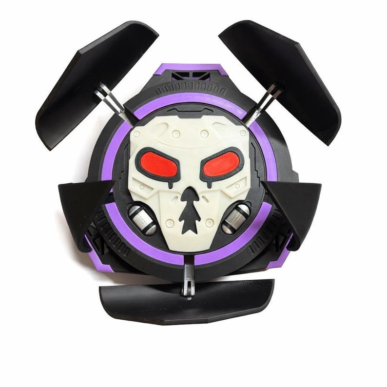 Puede incluir: Un accesorio en forma de calavera negro, morado y blanco con tres alas negras. El accesorio tiene un dise&ntilde;o circular con una cara de calavera en el centro, con cuencas rojas. Las alas est&aacute;n unidas con varillas plateadas.