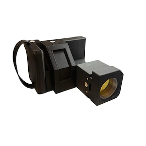 Op de afbeelding: Een zwart, rechthoekig apparaat met een lens en een riem. Het apparaat heeft een boxy ontwerp met een cilindrische lens. De lens heeft een gele binnenkant. De riem is zwart en aan de zijkant van het apparaat bevestigd.