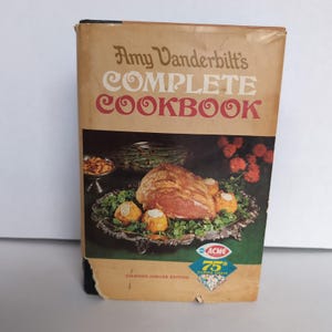 May include: A vintage cookbook titled "Amy Vanderbilt's Complete Cookbook" with a photo of a roasted meat dish on the cover. The book is a Diamond Jubilee Edition and features the Acme 75th Diamond Jubilee logo.