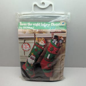May include: A Bernat Twas the night before Christmas craft kit in a plastic bag with a hanger. The package features two green and red stockings, one with a Santa Claus design and one with a candle and tree design, filled with small toys.