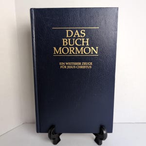 May include: A navy blue book with gold lettering that reads "DAS BUCH MORMON" and "EIN WEITERER ZEUG FÜR JESUS CHRISTUS". The book is standing upright on a black stand.