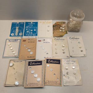May include: A collection of vintage mother of pearl buttons displayed on various branded cardboard backing cards, including Luckyday, Le Chic, American Maid, and Costumakers, alongside a clear glass jar filled with loose white buttons.