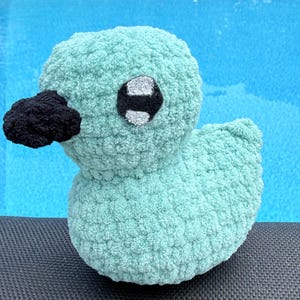 May include: A small, soft, crocheted duck toy made from light mint green yarn. The duck features a textured, plush surface, a solid black crocheted beak, and large, round eyes with black pupils and shimmering silver accents on the sides.