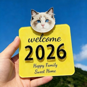 Puede incluir: Un letrero de bienvenida amarillo con el texto "welcome 2026 Happy Family Sweet Home". Una cabeza de gato con ojos azules y pelaje blanco y marrón está en la parte superior del letrero. El letrero se sostiene contra un cielo azul.