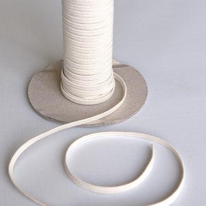 Pode incluir: Uma bobina de cordão de algodão branco, enrolada em torno de um tubo de cartão, com um comprimento de cordão enrolado em uma superfície branca.
