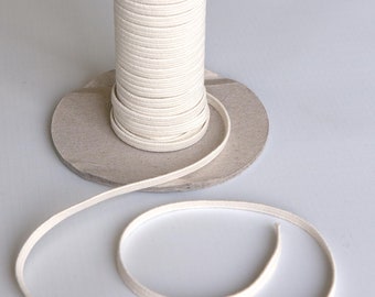 Puede incluir: Una bobina de cordón de algodón blanco, enrollada alrededor de un tubo de cartón, con una longitud de cordón enrollada sobre una superficie blanca.