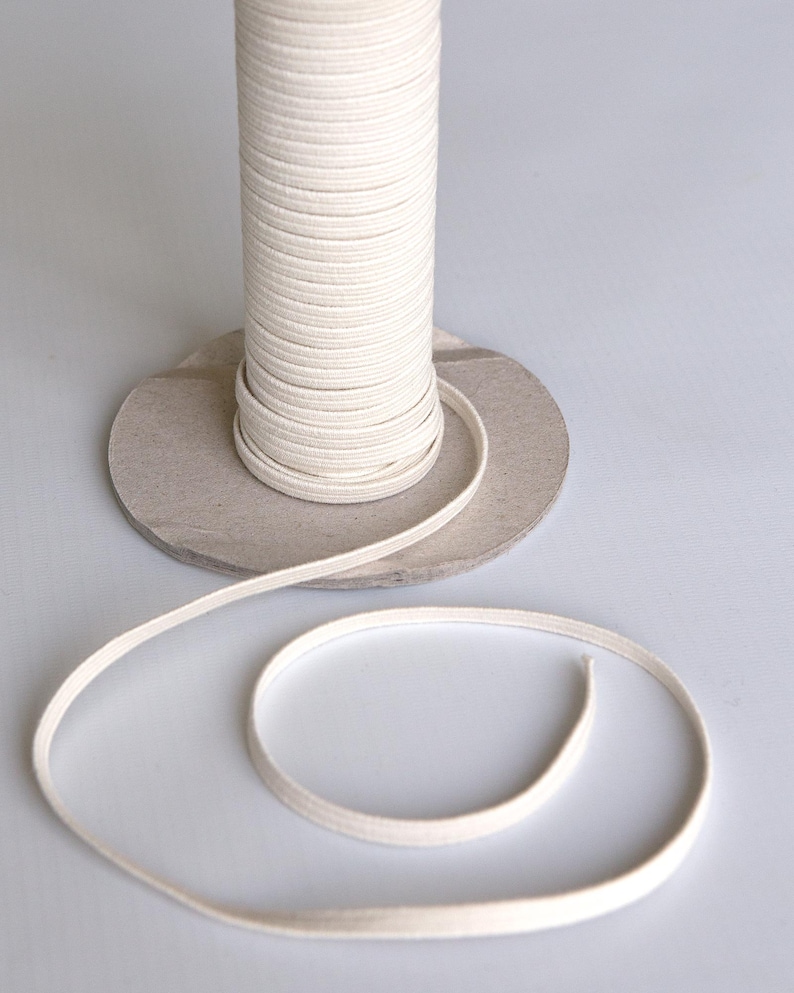 May include: A spool of white cotton cord, wound around a cardboard tube, with a length of cord coiled on a white surface.