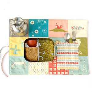 Peut inclure: Un set de table patchwork coloré avec une gourde en acier inoxydable, une boîte à lunch divisée contenant une tomate, du pain et des raisins, et une poche contenant une cuillère et une fourchette. Le set de table présente des illustrations fantaisistes.