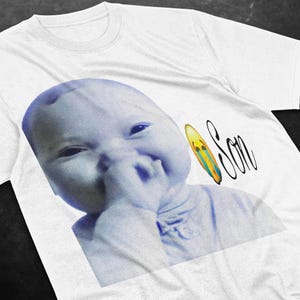May include: White t-shirt featuring a blue-toned image of a baby with a hand near its mouth. A yellow, blue, and green surfboard graphic is next to the word "Son" in a cursive font.