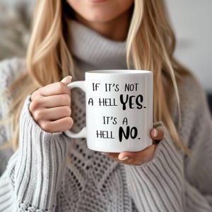 May include: White ceramic mug with the text "IF IT'S NOT A HELL YES. IT'S A HELL NO." in black. The mug has a white handle and is held by a person wearing a gray sweater.
