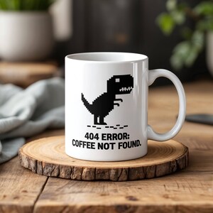 Może przedstawiać: Biały ceramiczny kubek do kawy z czarnym pikselowanym dinozaurem i tekstem "404 ERROR: COFFEE NOT FOUND."