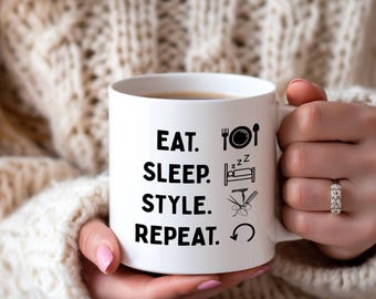 Puede incluir: Taza de cerámica blanca con las palabras "EAT. SLEEP. STYLE. REPEAT." en negro, junto con iconos. La taza es sostenida por una persona que lleva un suéter color crema.