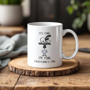 Könnte beinhalten: Weiße Keramik-Tasse mit einem schwarz-weißen Cartoon-Vogel-Design. Auf der Tasse steht der Text "It's fine. I'm fine. Everything's fine.". Die Tasse steht auf einem Holzuntersetzer.