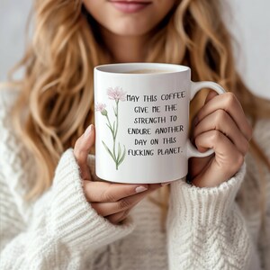 May include: White ceramic coffee mug with a floral design and the text "MAY THIS COFFEE GIVE ME THE STRENGTH TO ENDURE ANOTHER DAY ON THIS FUCKING PLANET." The mug has a curved handle and is filled with coffee.