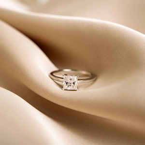 May include: A silver ring with a square-shaped diamond on a beige fabric background.
