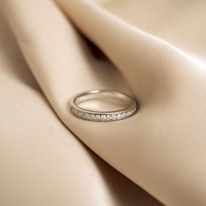 Może przedstawiać: Delikatny srebrny pierścionek z paskiem ozdobionym rzędem małych, czystych diamentów osadzonych w gładkiej, wypolerowanej obrączce.