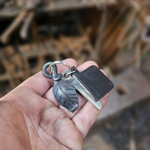 Op de afbeelding: Een zilveren sleutelhanger met een zwarte sleutelhouder en een bladvormige hanger. De hanger is van metaal en heeft een rustieke afwerking.