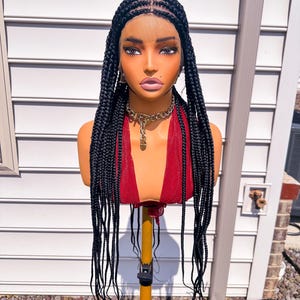 May include: A black braided wig with long, straight, individual box braids styled with a centre part. The wig features a sheer lace front base and is displayed on a mannequin head wearing a deep red sleeveless top and a gold chain necklace.
