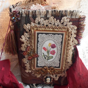May include: A burgundy fabric-covered journal with a gold-trimmed frame featuring a floral design with tulips. The journal is decorated with lace, beads, and a butterfly embellishment.