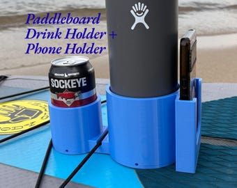 Op de afbeelding: Een lichtblauwe kunststof bekerhouder en telefoonhouder voor paddleboards, voorzien van een cilindrische bekerhouder, een flessenhouder en een verticale gleuf voor een smartphone. De tekst Paddleboard Drink Holder + Phone Holder is zichtbaar.