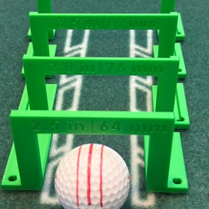 May include: A white golf ball with red stripes is positioned beneath four green, rectangular measuring guides. The guides are labeled with measurements in inches and millimeters, ranging from 2.5 in (64 mm) to 4.0 in (102 mm).