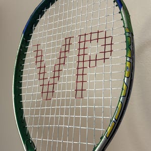 May include: A tennis racket with a green and silver frame featuring a white string bed. The strings are marked with a large red letter V and a red letter P. A small black rectangular label at the base of the string bed reads SOFT SHOCK.