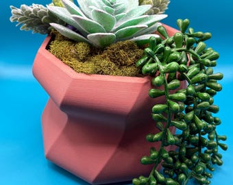 May include: A pink geometric planter with a succulent plant and trailing greenery. The planter has a unique, faceted design.