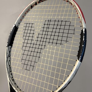May include: A Wilson Hammer tennis racket featuring a white, black, and red frame with an oversized head. The strings are white with a prominent black stencil of the letter P in the center. The frame includes the text Hammer and Carbon Matrix.