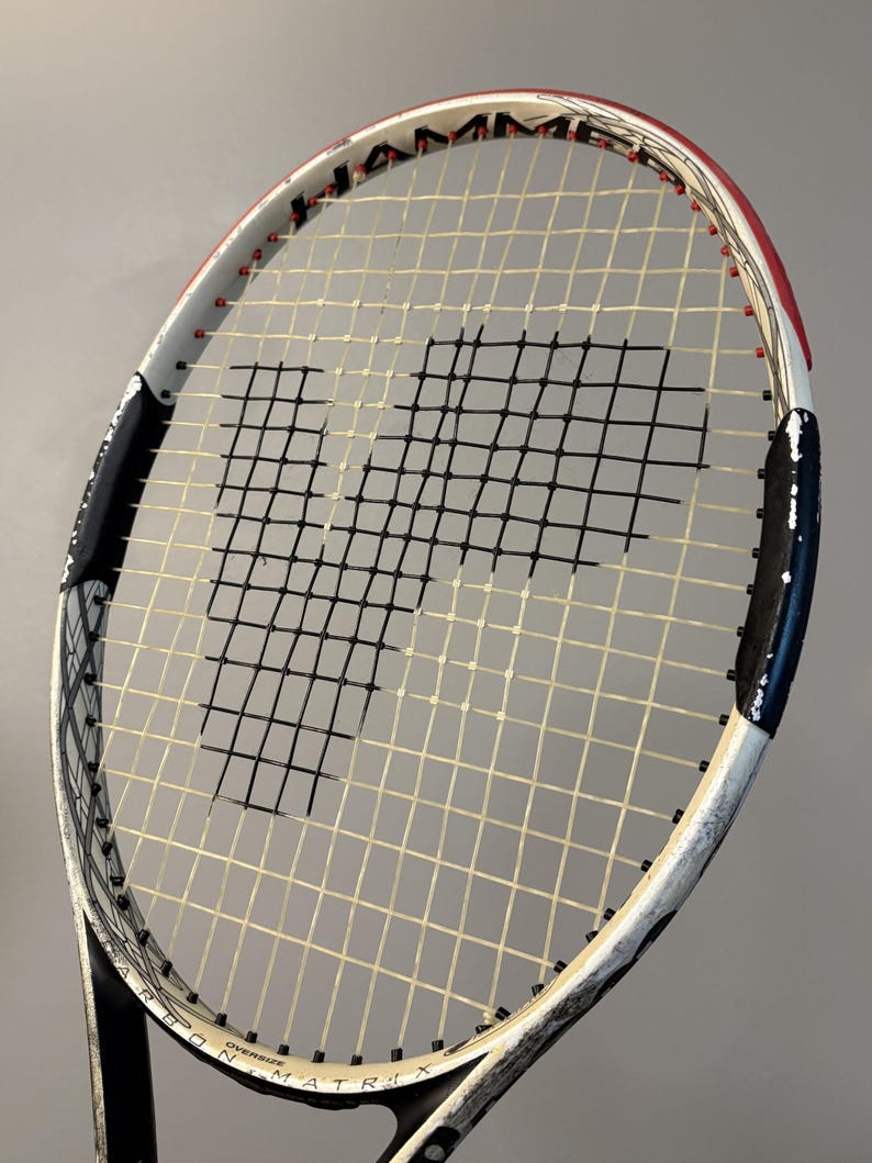 May include: A Wilson Hammer tennis racket featuring a white, black, and red frame with an oversized head. The strings are white with a prominent black stencil of the letter P in the center. The frame includes the text Hammer and Carbon Matrix.