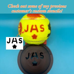 May include: A yellow tennis ball featuring the red printed text JAS and a star icon, positioned above a dark brown circular stencil tool with the same JAS text and star cutout. The image includes the text Check out some of my previous customer's custom stencils!