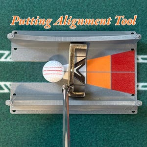 May include: A gray plastic putting alignment tool with the text Putting Alignment Tool printed above it. The device features a central channel, a white golf ball with red stripes, and a putter head positioned over orange and red grid markings.