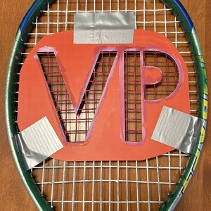 May include: A green and blue tennis racket featuring a large orange rectangular insert with the letters VP cut out. The orange insert is secured to the white racket strings with three pieces of silver duct tape. The frame is labeled Titanium.