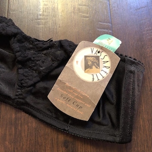 Pode incluir: Um sutiã preto de copa macia com detalhes em renda na parte superior. Uma etiqueta de cartão castanha está presa, exibindo a marca Vassarette, uma ilustração de um busto clássico e o texto: soft cup bra provides easy support and a gentle, natural look.