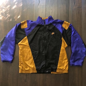 May include: A long-sleeve zip-up jacket featuring a color-blocked design in black, purple, and gold. The jacket has a high collar, elastic cuffs, and a hemline. A small orange Nike logo is embroidered on the upper left chest area.