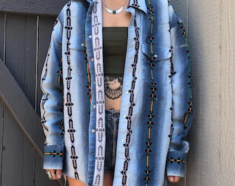 Puede incluir: Una camisa azul claro con botones y un diseño de inspiración occidental. La camisa presenta un patrón vertical de formas geométricas negras, naranjas y verdes. La camisa tiene dos bolsillos en el pecho y una tira estampada blanca y gris en el centro.