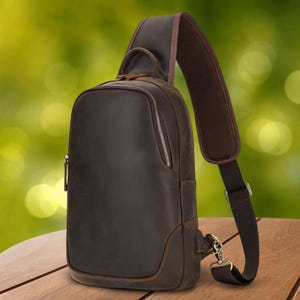 May include: A brown leather sling bag with a zippered main compartment and a padded shoulder strap. The bag has a small handle at the top and a metal clasp for the strap. The bag is sitting on a wooden surface.