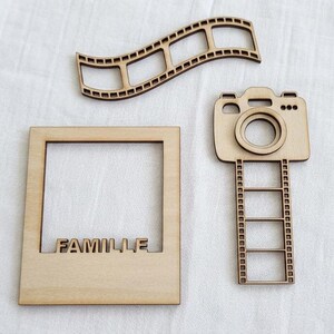 May include: Three light-colored wooden cutouts featuring a wavy film strip, a square photo frame with the word FAMILLE cut out at the bottom, and a camera shape attached to a vertical film strip with four rectangular openings for photos.