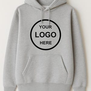 May include: A light gray hoodie with a front pocket and a hood. The front of the hoodie features a black circle with the text "YOUR LOGO HERE" in white. The hoodie is made of a soft material.