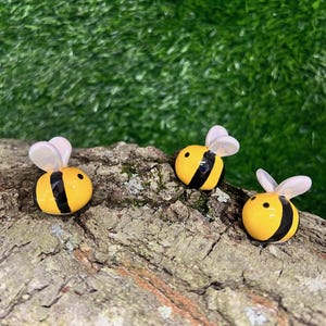 May include: Three small, yellow bee figurines with black stripes and translucent white wings rest on a piece of wood. The bees have a glossy finish and are set against a blurred green background.