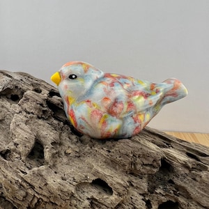 May include: A ceramic bird figurine with a colorful, marbled design. The bird features a yellow beak, black eyes, and a mix of red, yellow, and blue hues. The figurine is resting on a piece of weathered wood.