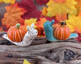 May include: Two ceramic snail figurines, one with a white body featuring black speckles and the other with a solid teal body. Each snail carries a glossy orange pumpkin on its back with a small green or brown stem, resting on a piece of driftwood.