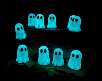 May include: A collection of ten small, glowing, light blue ghost figurines, each featuring two dark, oval-shaped eyes. The figures are arranged in two rows against a dark background, emitting a soft, luminous light from their bodies.