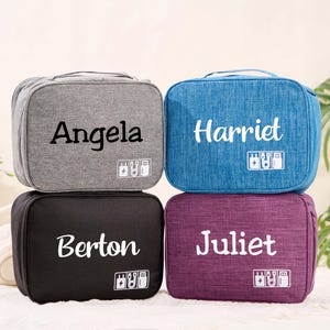 Peut inclure: Quatre trousses de voyage personnalisées en gris, bleu, noir et violet. Chaque trousse porte un nom imprimé en blanc, avec un petit graphique d'appareils électroniques. Les trousses sont rectangulaires avec une poignée.