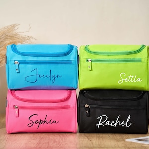 Peut inclure: Quatre trousses de voyage personnalisées en bleu, vert, rose et noir. Chaque sac a une fermeture éclair et une tirette assortie. Les noms "Jocelyn", "Settla", "Sophia" et "Rachel" sont écrits en cursive sur le devant.