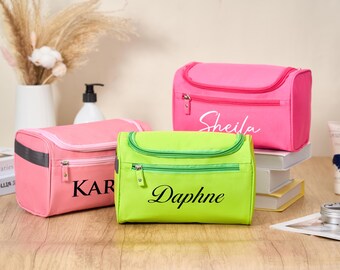 May include: Three personalized cosmetic bags in pink and green. The pink bags have the names "Sheila" and "Kar" printed on them. The green bag has the name "Daphne" printed on it. Each bag has a zipper closure.