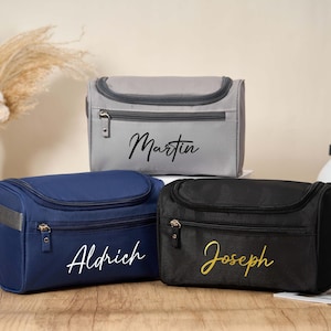 Könnte beinhalten: Drei personalisierte Kulturbeutel in Grau, Marineblau und Schwarz. Jede Tasche hat einen Reißverschluss und eine gewölbte Oberseite. Die Namen "Martin", "Aldrich" und "Joseph" sind in Schreibschrift auf die Vorderseite geschrieben. Die Taschen stehen auf einer Holzoberfläche.